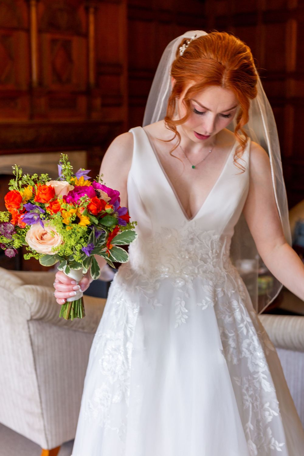 Bride in white lace wedding dress holds vibrant multicolored bouquet with roses and greenery in elegant indoor setting.