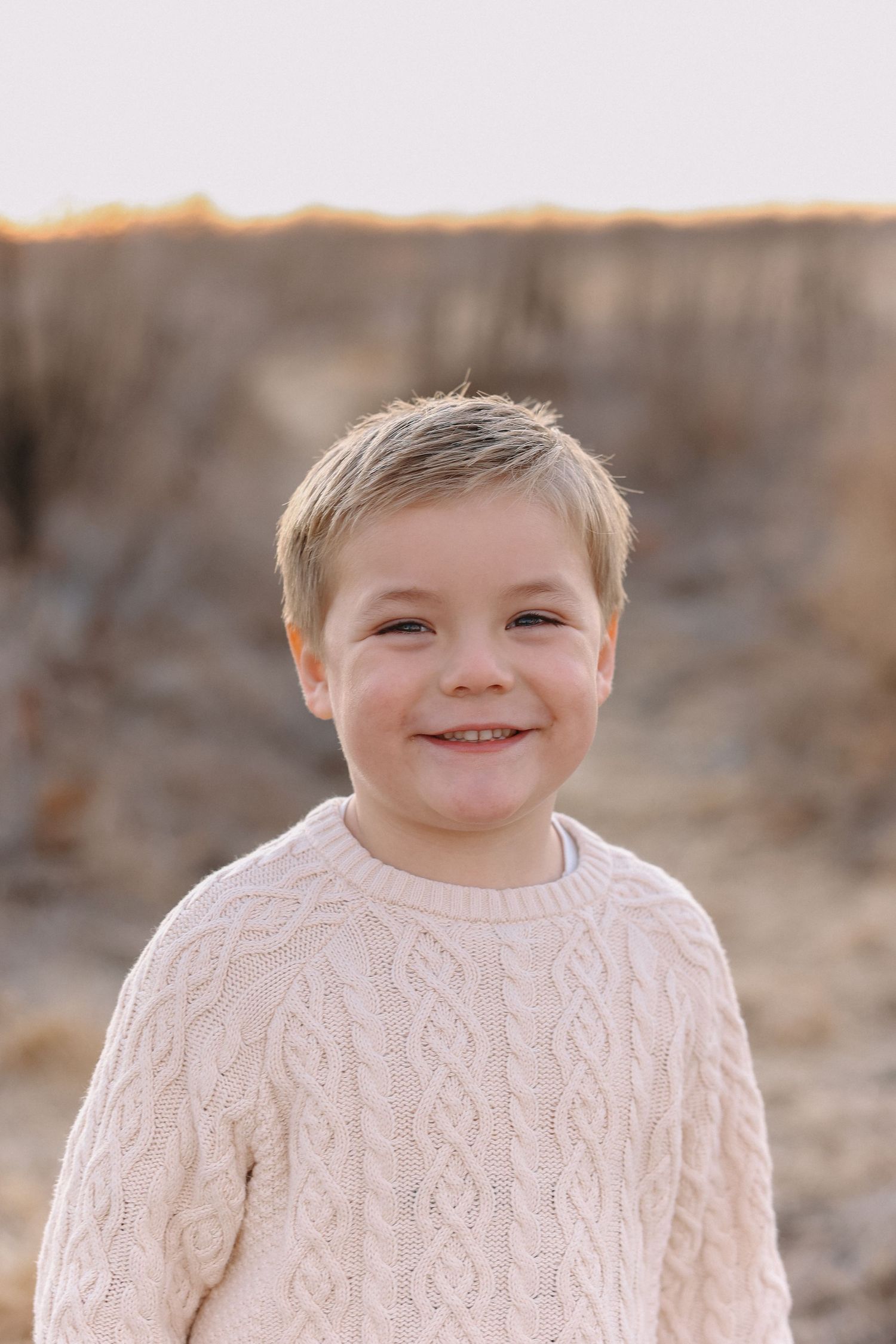 Child wearing cream cable knit sweater smiles warmly in outdoor portrait series.