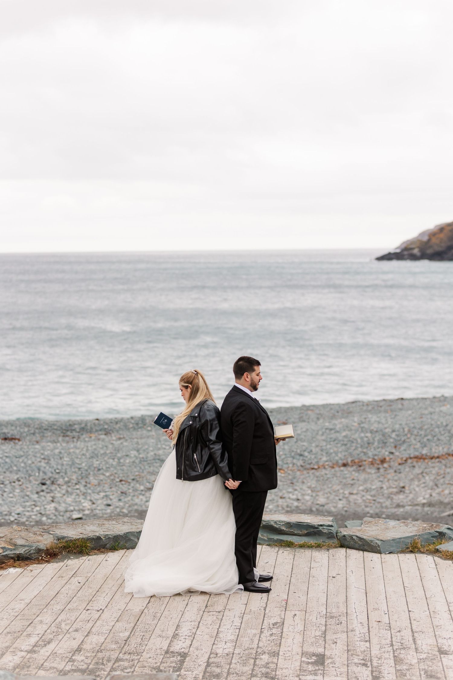 A couple in wedding attire walks along a wooden boardwalk by the ocean on an overcast day.