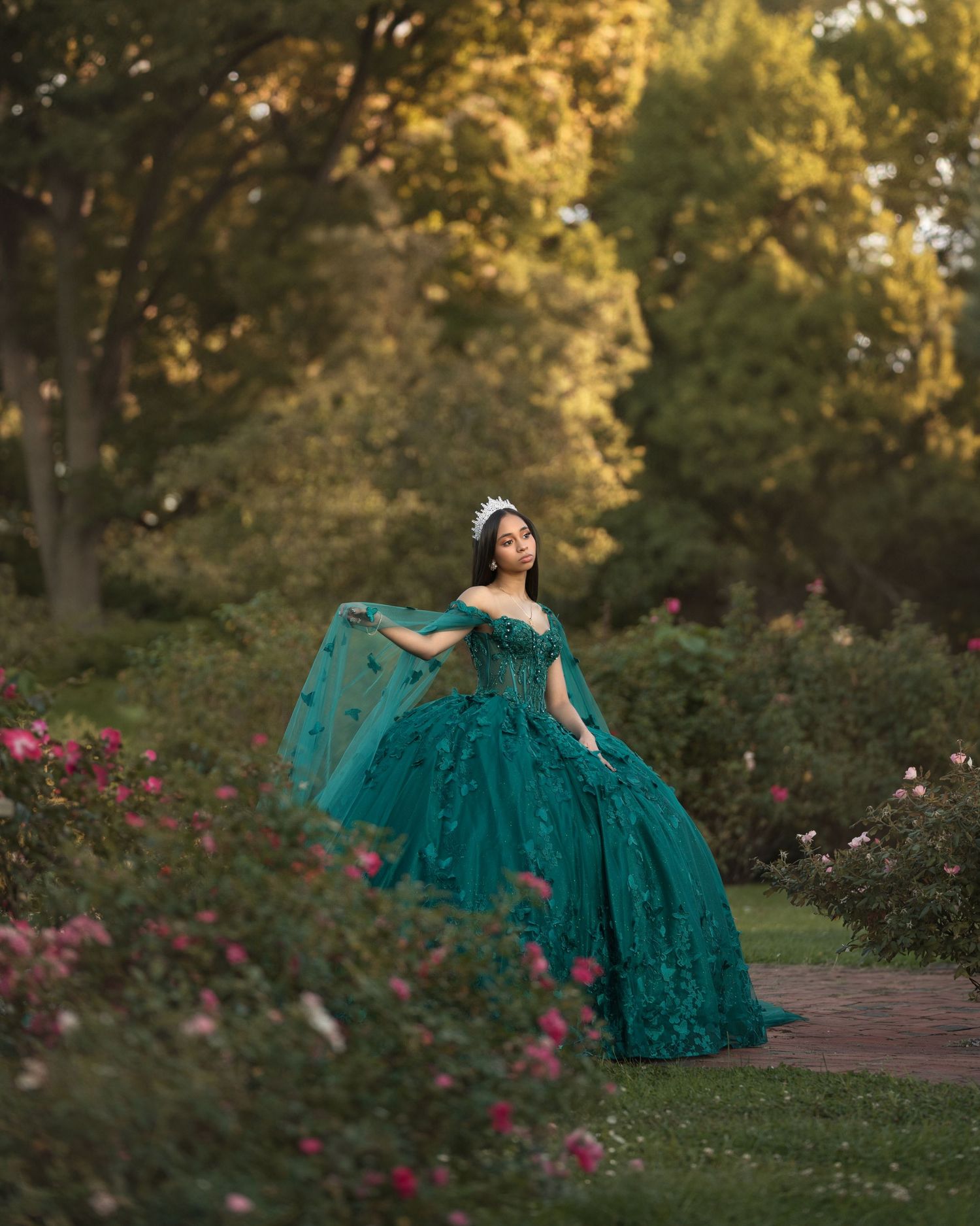 A flowing emerald green ballgown dress twirls gracefully in a garden setting with pink flowers and trees at sunset.