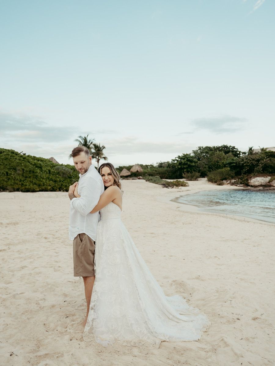 A romantic beach wedding couple embraces on sandy shores with flowing white dress and ocean backdrop.