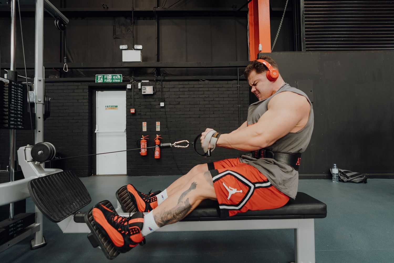 Muscular person in orange shorts performing a rowing exercise on exercise equipment in a dark gym setting.