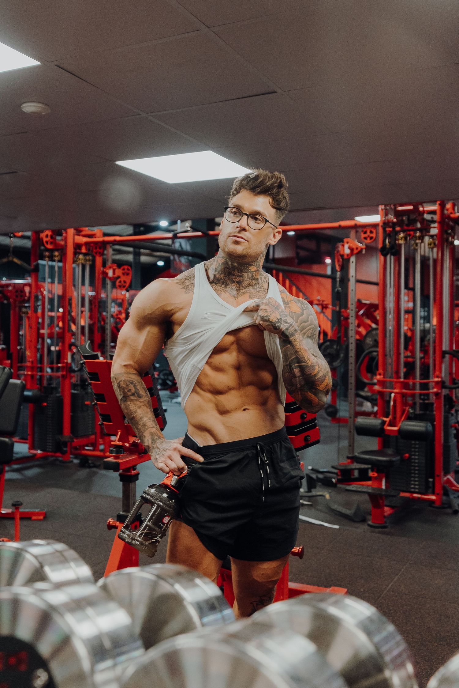 Tattooed fitness model in tank top displaying muscles in red-equipped gym.