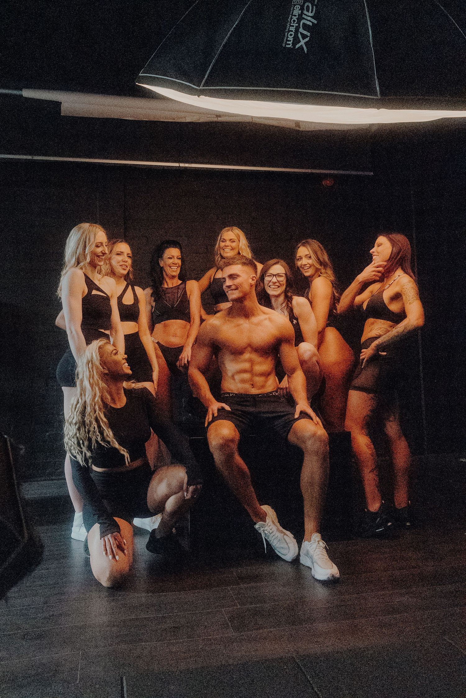 A group poses together in a dark studio setting with dramatic lighting and fashion-forward styling.