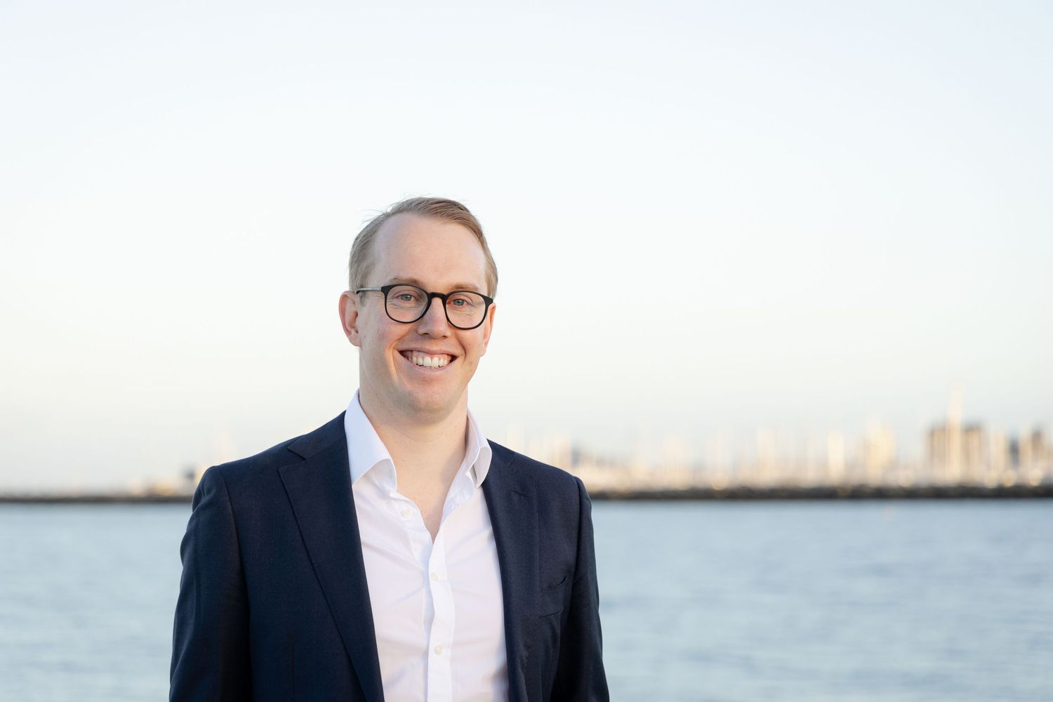 A person in a navy suit and white shirt smiles while standing near a waterfront with buildings visible in the distance.