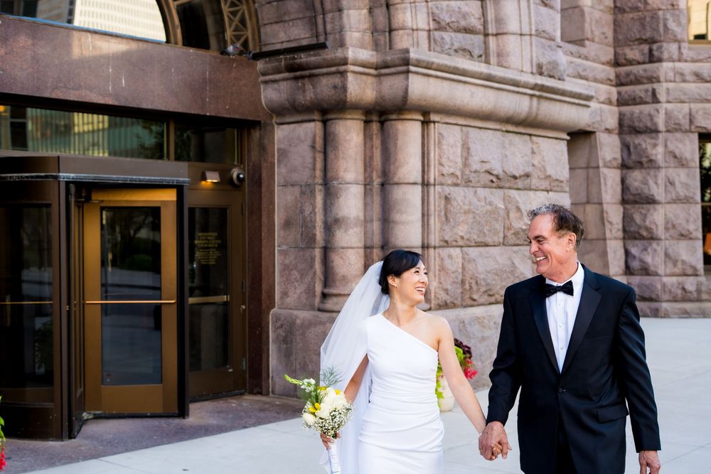 A couple in formal wedding attire walking and smiling together outside a stone building.