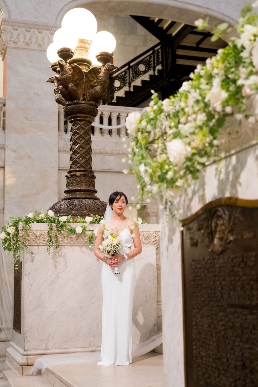 Elegant bride in white dress holds bouquet while standing in ornate marble colonnade with decorative pillars.