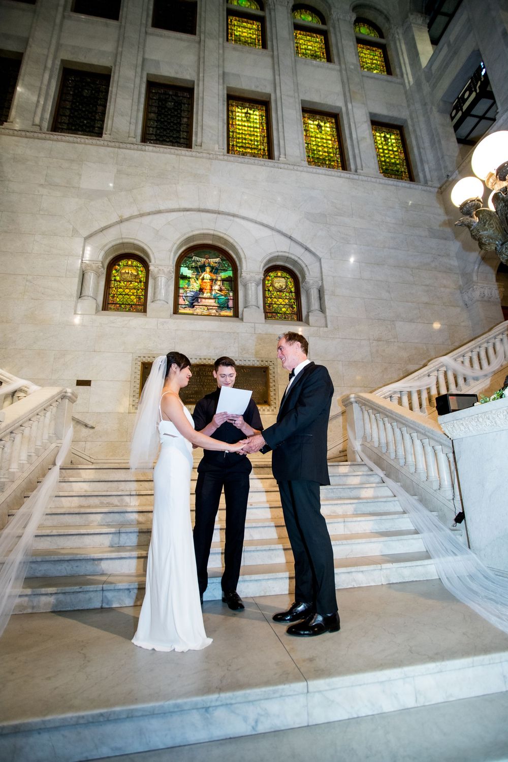 Wedding ceremony taking place on grand marble staircase with stained glass windows.