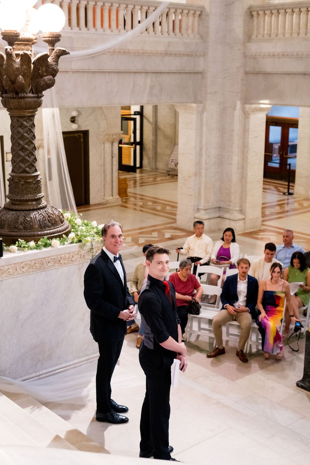Wedding ceremony taking place in ornate marble interior with columns and gathered guests.