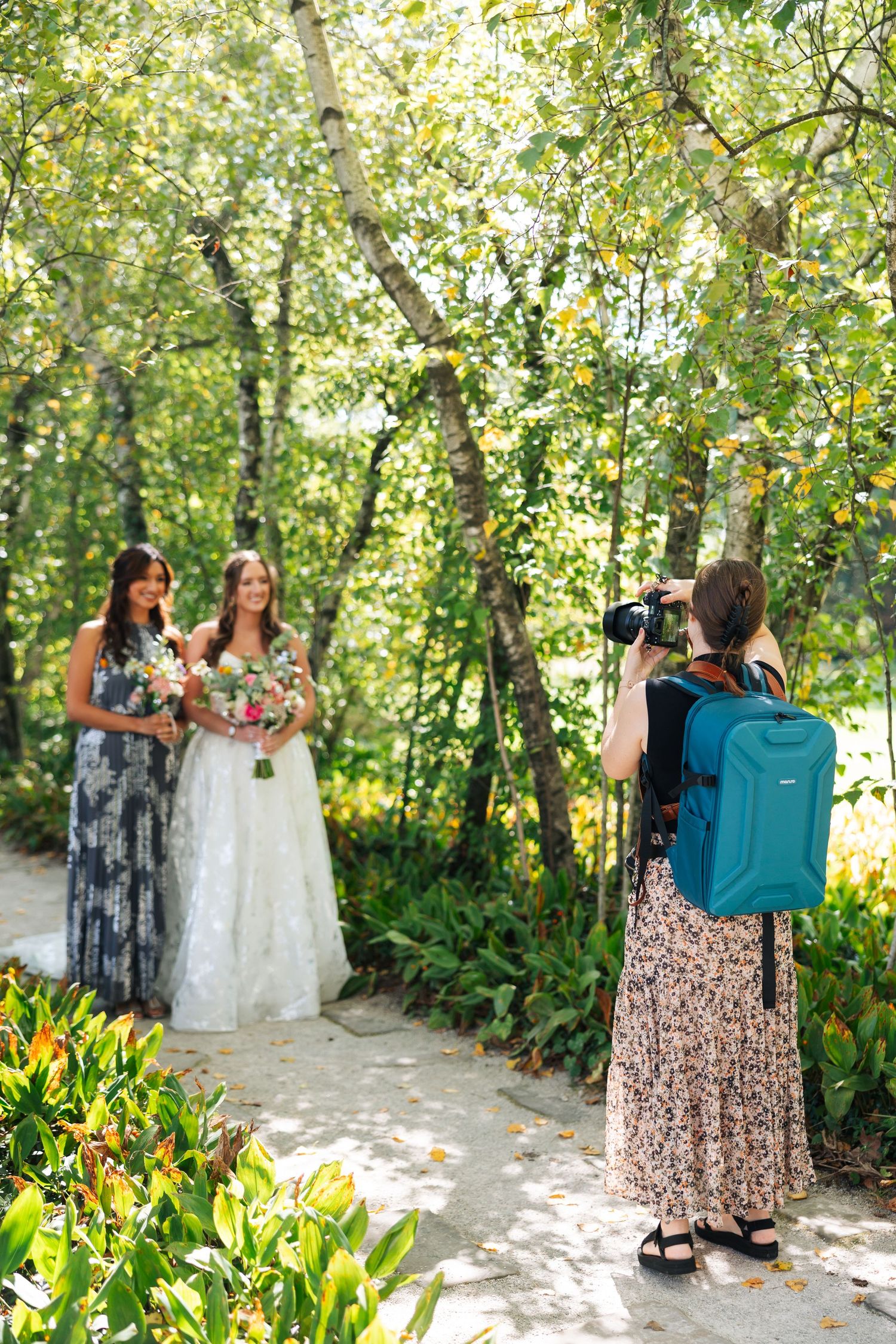 A photographer with a blue backpack captures a wedding scene on a garden path surrounded by lush greenery.