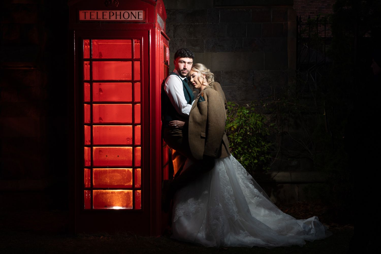 A couple shares a romantic moment in front of an illuminated red British phone booth at night.