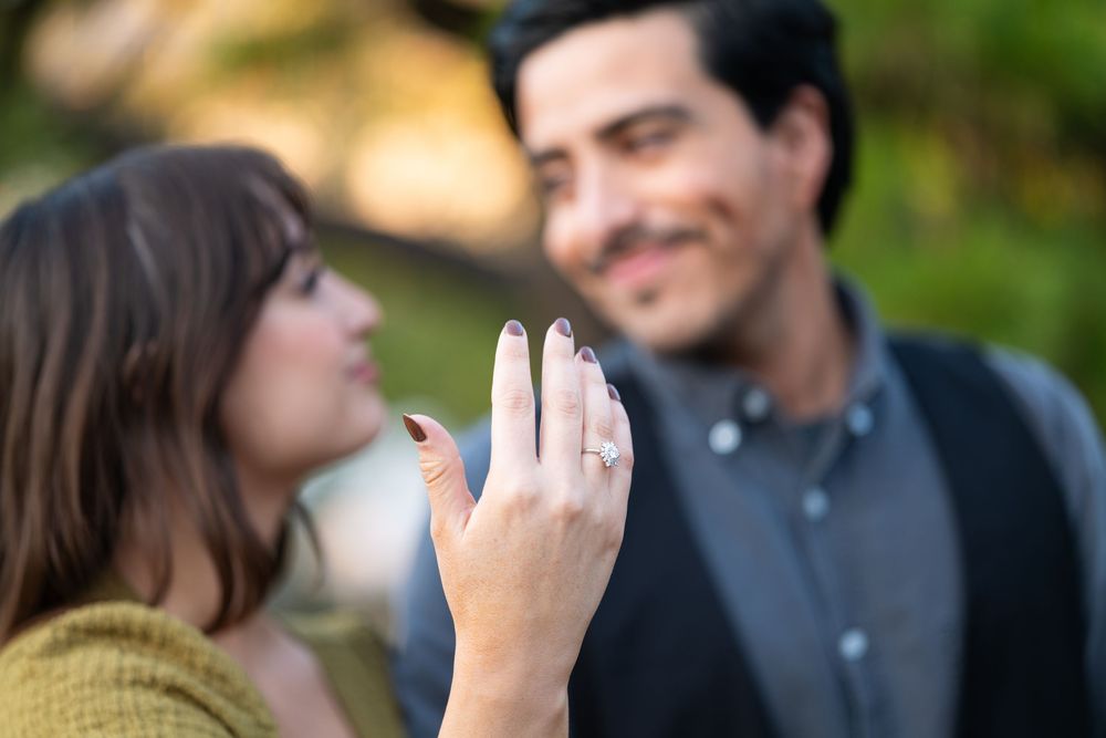 Woman proudly displaying engagement ring with smiling fiance in soft bokeh background.
