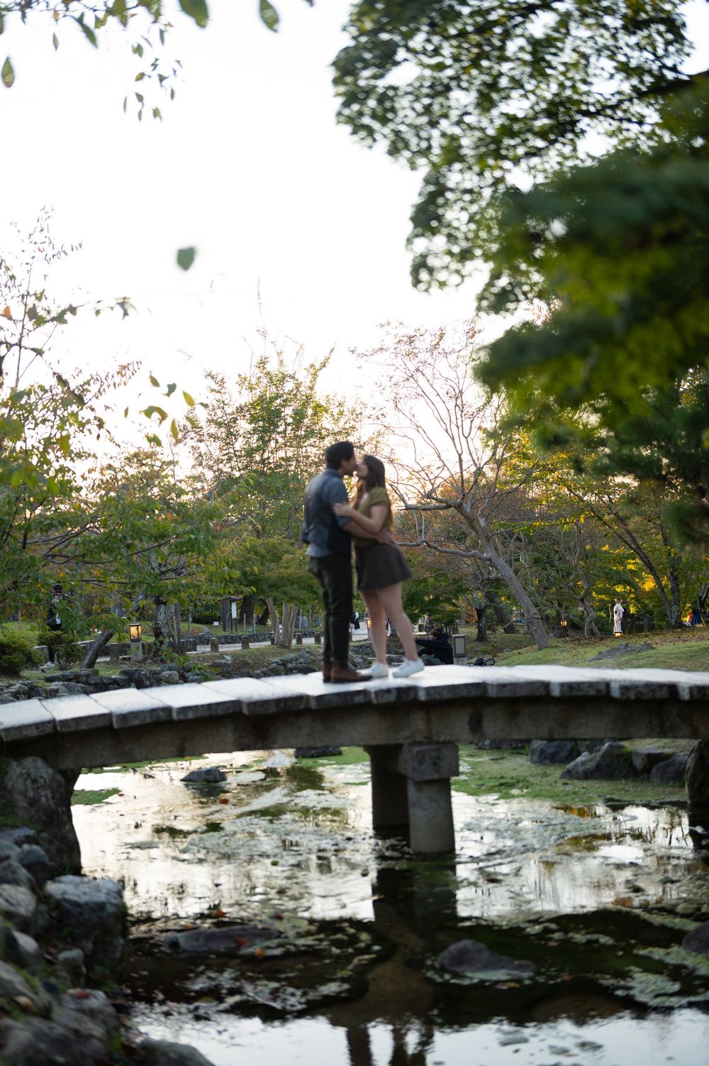 Couple sharing a romantic kiss on a stone arch bridge over a reflective pond in a lush Japanese garden.