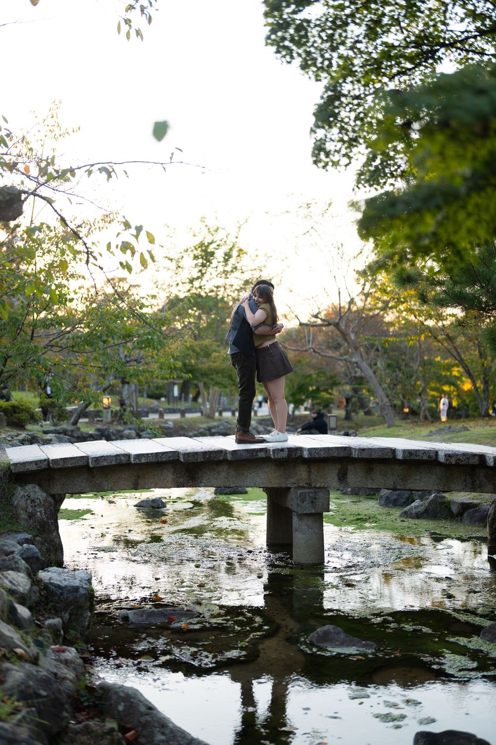 Couple embracing on a curved stone bridge surrounded by serene Japanese garden greenery and calm water.