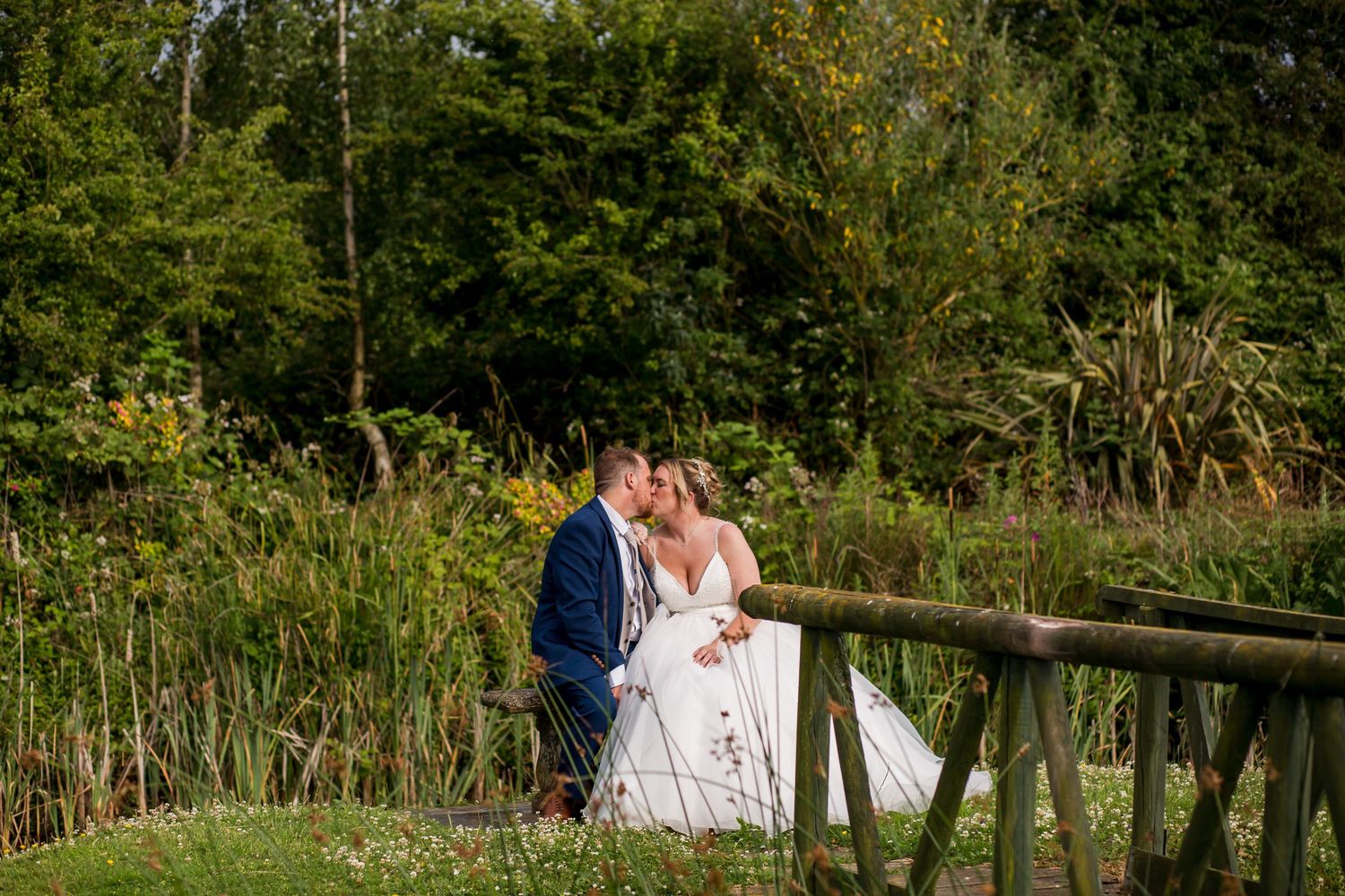 Newlyweds walk along a rustic wooden fence surrounded by lush summer foliage.