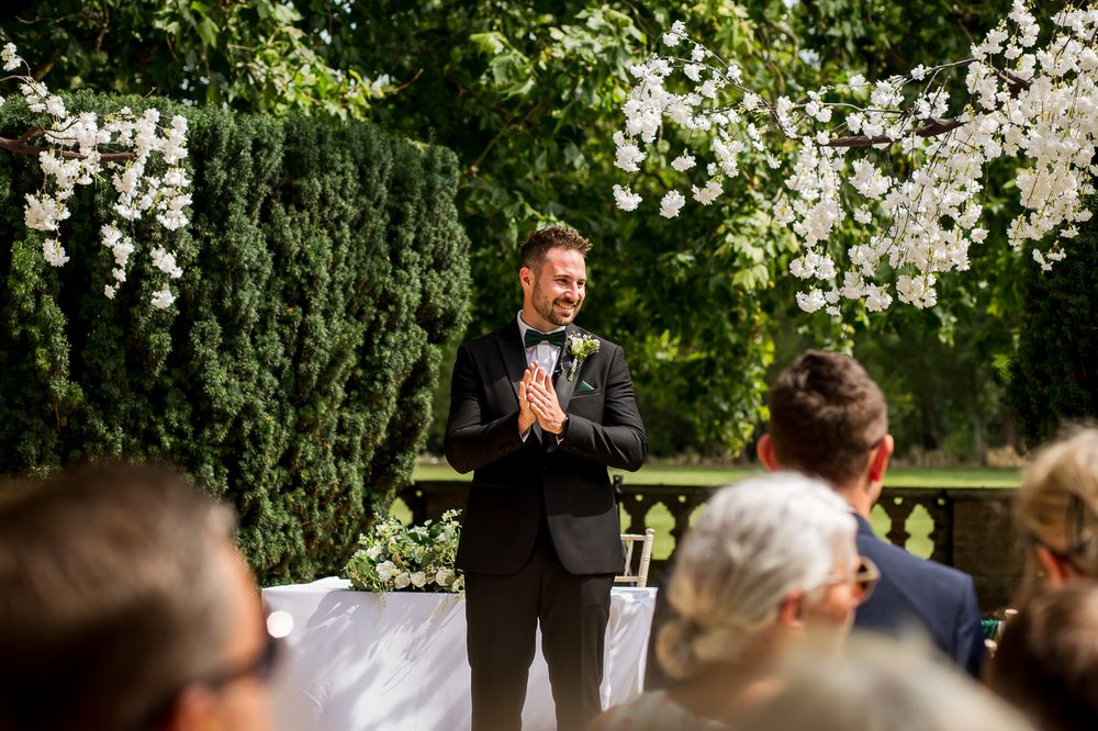 Groomsmen laugh together during a wedding ceremony in a scenic garden setting.