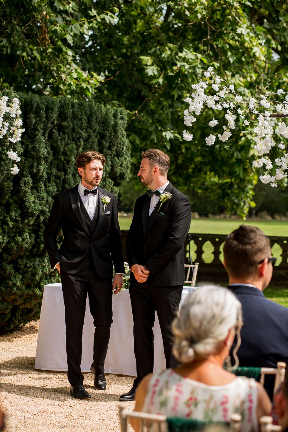 Wedding party members interact during an outdoor ceremony with guests seated in the background.