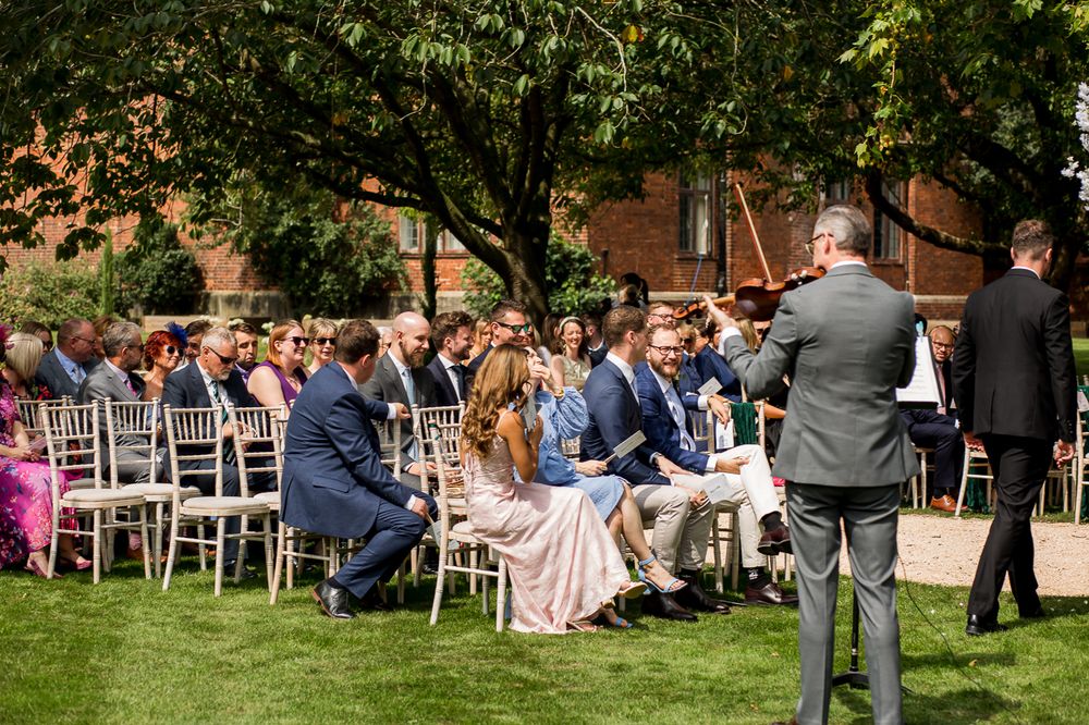 Wedding guests seated in white chairs during an outdoor ceremony near a brick building.