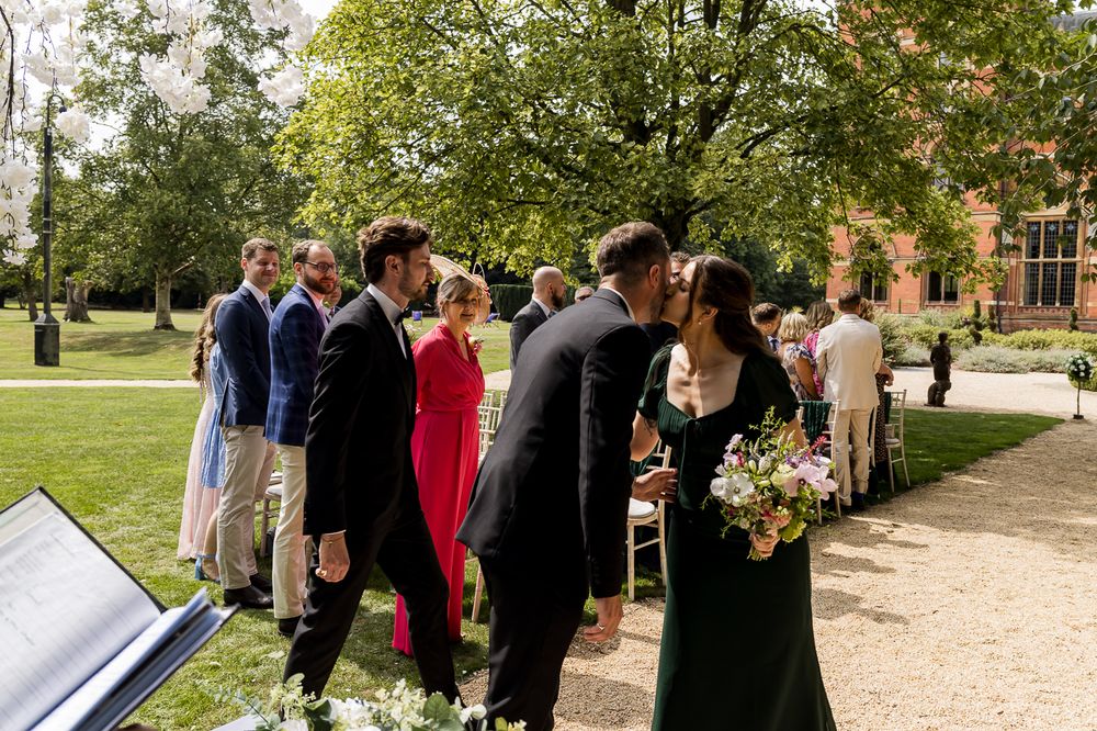Wedding guests gathered outside a Gothic revival building with manicured gardens and pathways.