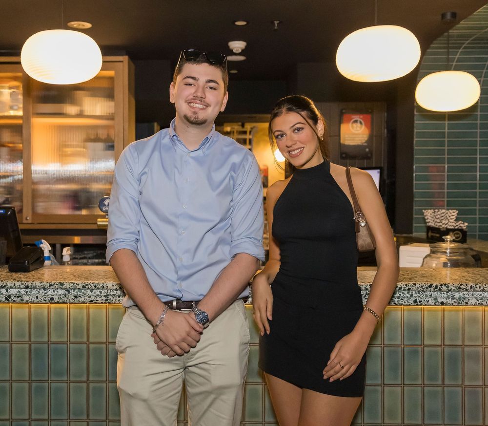 People in casual attire stand together smiling in front of a restaurant counter with pendant lights overhead.