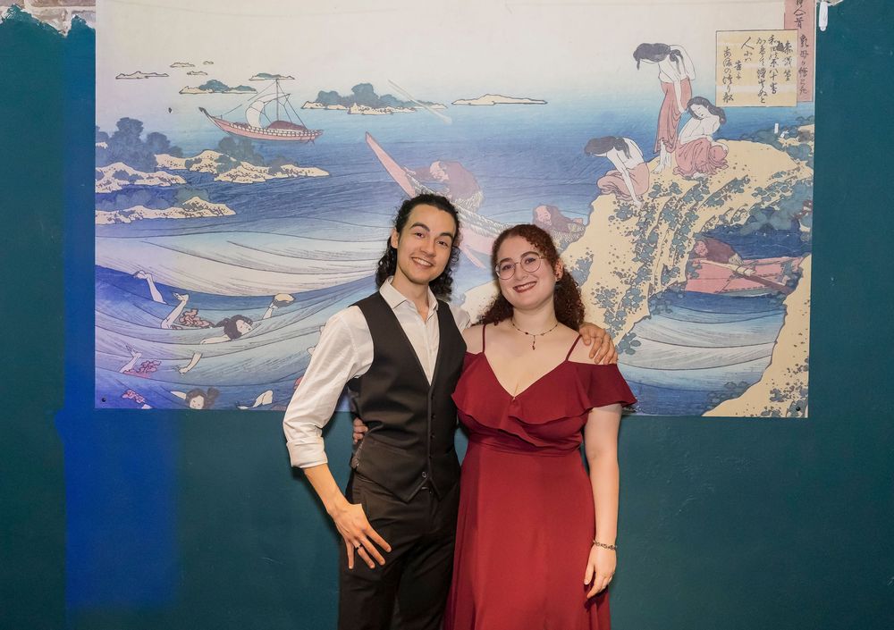 Two people in formal attire pose in front of a Japanese art mural featuring a coastal landscape with boats.