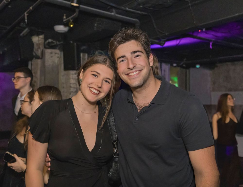Two people in dark clothing smile together at a nightclub with purple lighting in the background.