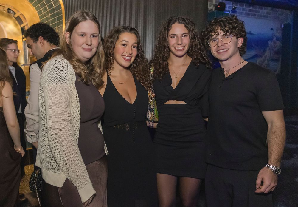 A group of young people pose together at a social gathering, wearing casual and dressy outfits in dark colors.