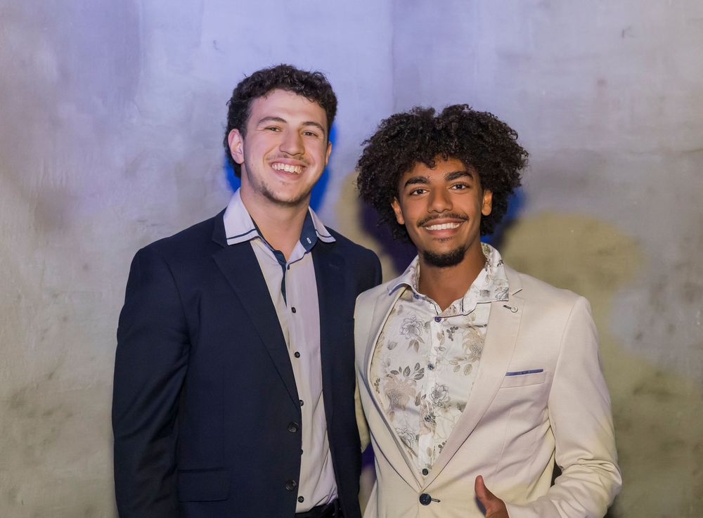 A group poses together at a formal event wearing suits and smiles against a textured wall background.