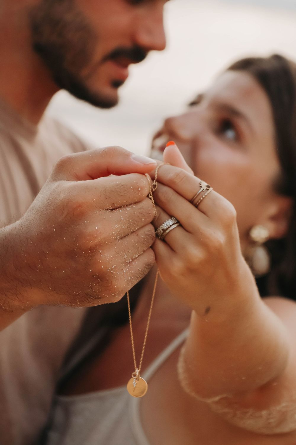 Close-up of couple holding hands with gold necklace, woman wearing stacked rings, sandy beach background.