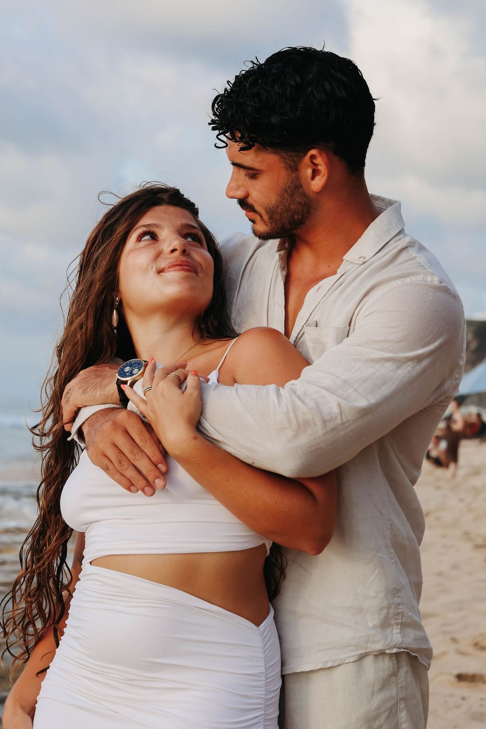 Couple embracing on beach, woman in white crop top and skirt smiling up at man in white shirt.