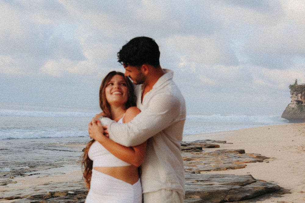 Couple hugging on beach with cloudy sky, woman in white crop top smiling up at man in white shirt.