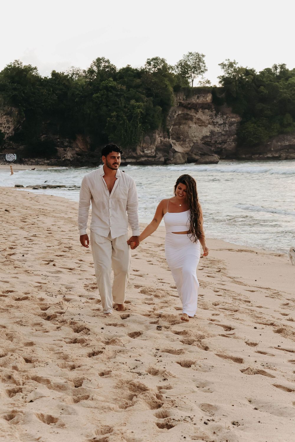 Couple in white outfits walking hand-in-hand on sandy beach with rocky cliffs and ocean in background.