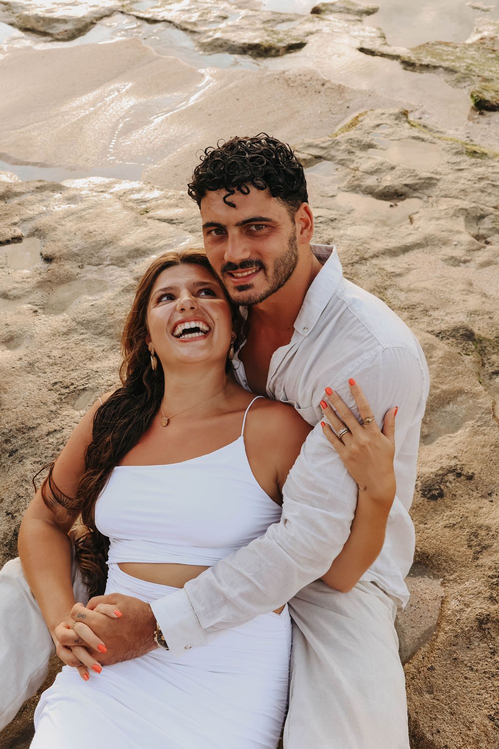 Laughing couple sitting on sandy beach, woman in white dress embraced by man in white linen shirt.
