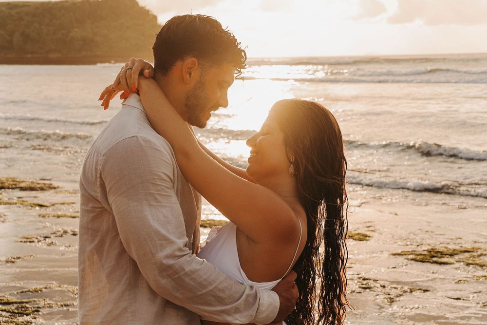 Couple facing each other in romantic embrace on beach during golden sunset with ocean waves behind them.