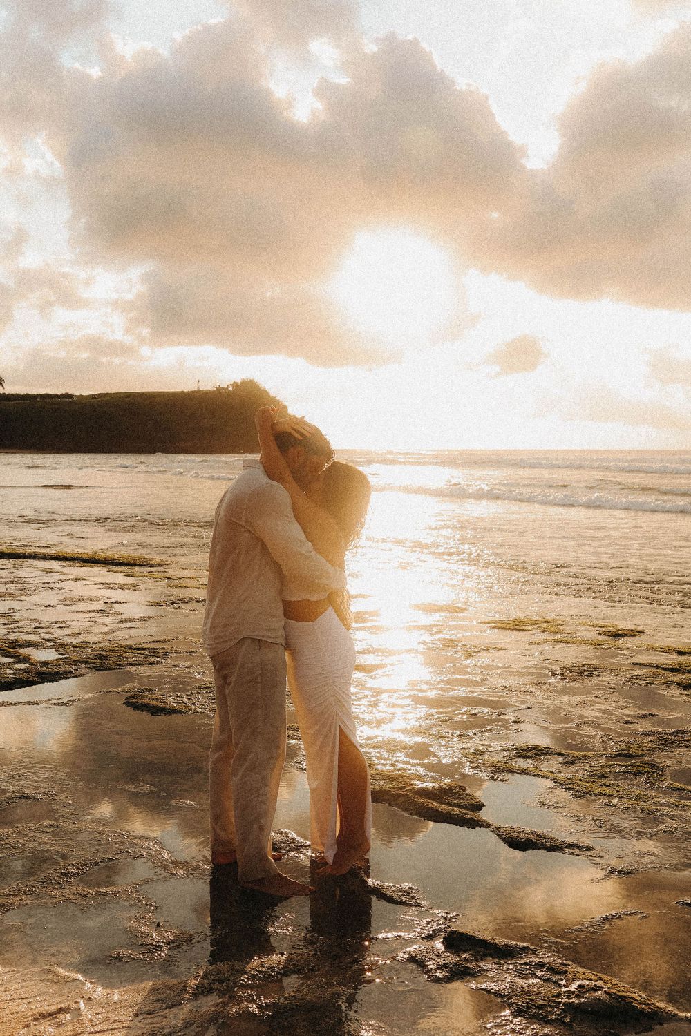 Romantic couple kissing at sunset on a reflective beach with dramatic golden sky and ocean backdrop.