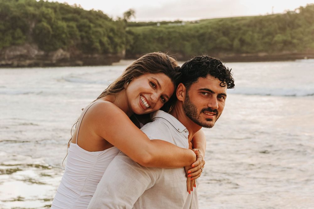 Couple embracing on a tropical beach, woman smiling and hugging man from behind with ocean waves in background.