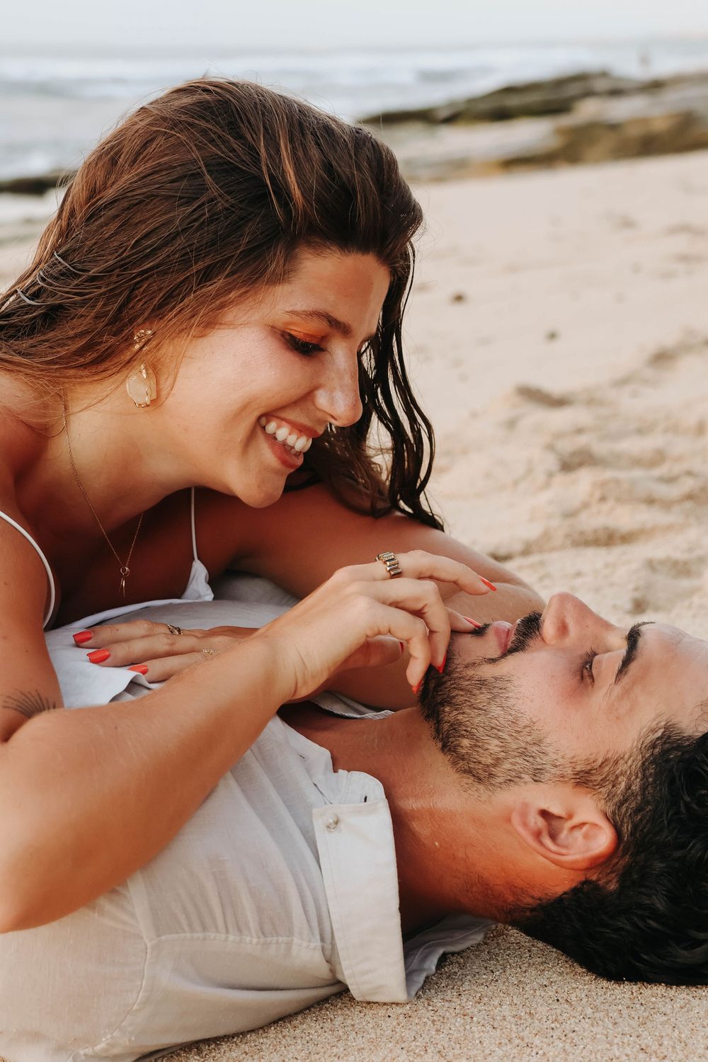 Woman smiling and lying on man on sandy beach, sharing a playful romantic moment outdoors.