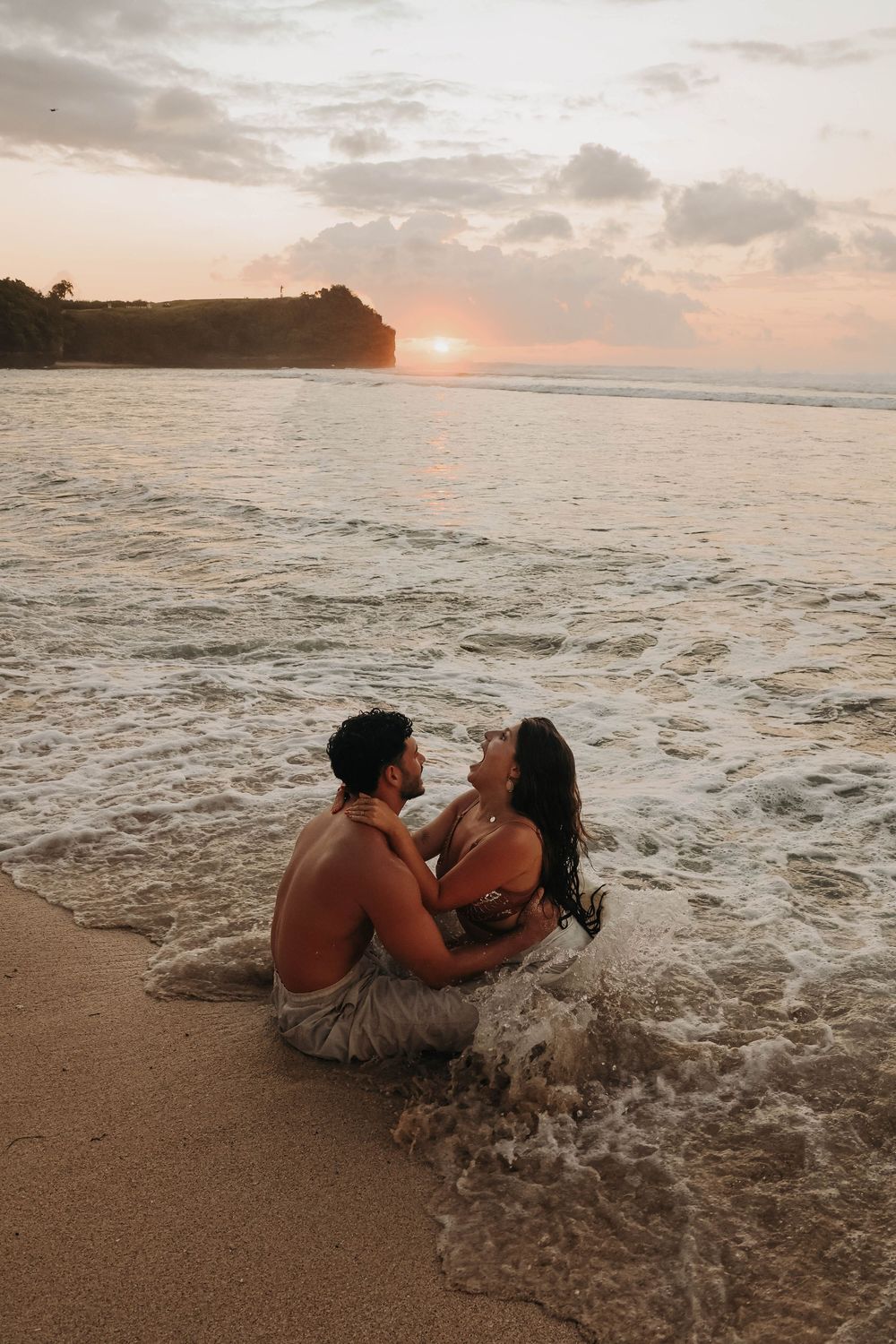 Couple sitting together in ocean waves at sunset on a tropical beach, sharing an intimate moment.