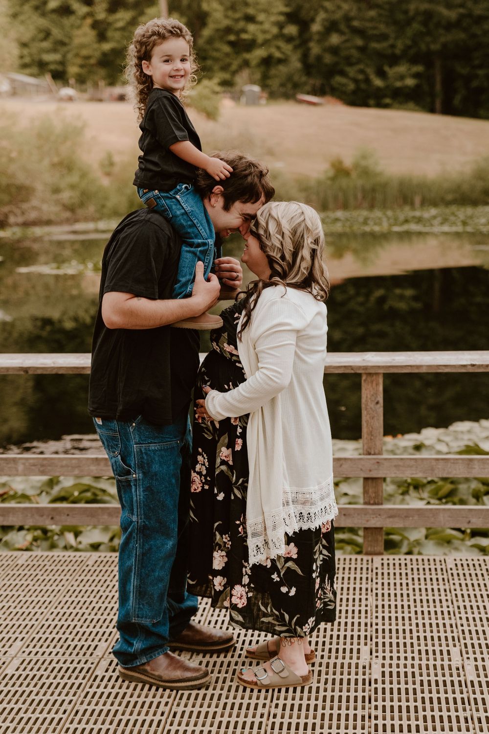 Happy family with toddler on dad's shoulders sharing a kiss near a scenic pond.