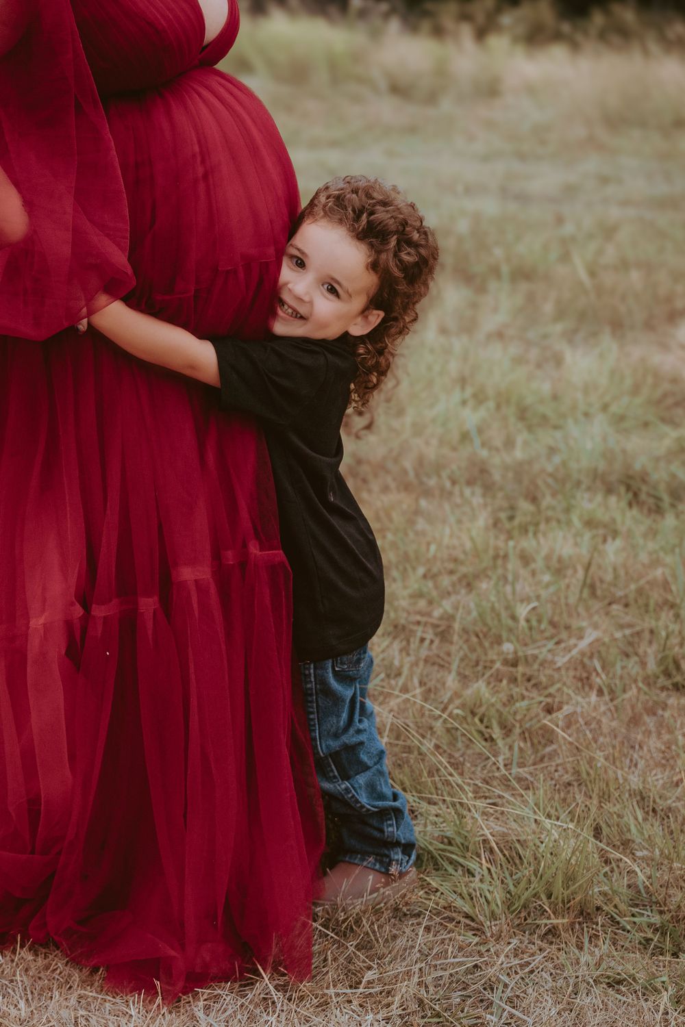 Curly-haired child hugging pregnant mother's belly in red tulle dress in a field.