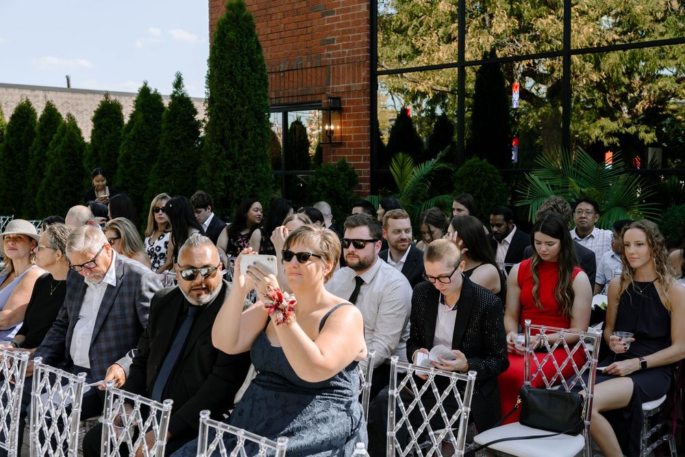 Wedding guests gather outdoors in front of a brick building for a ceremony on a sunny autumn day.