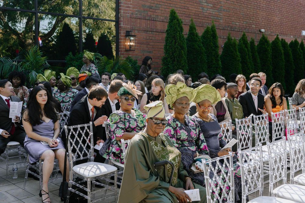 Wedding guests seated in white lattice chairs at an outdoor ceremony venue with brick wall and greenery backdrop.