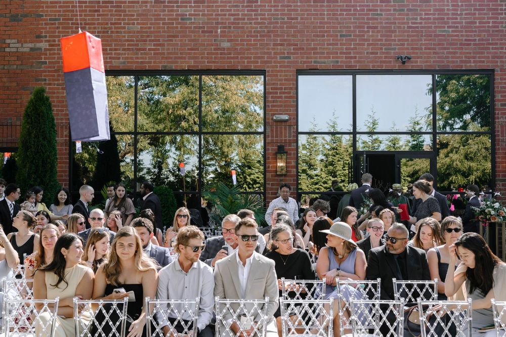 Wedding guests seated in white chairs at an outdoor ceremony venue with brick wall and greenery backdrop.