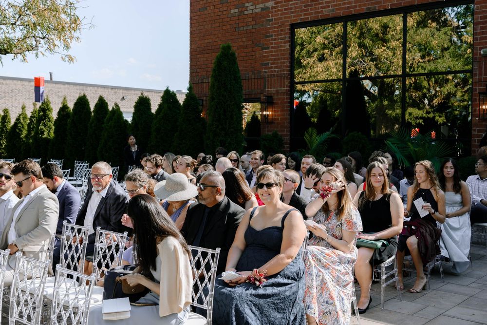 Wedding guests enjoy refreshments and conversation at an outdoor reception with white chairs and brick building backdrop.