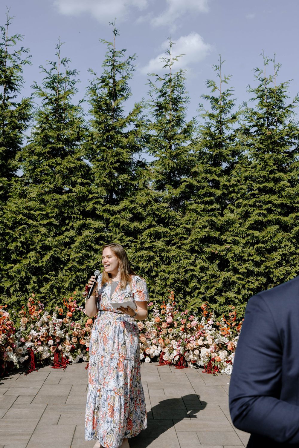 A speaker in a floral dress addresses guests at an outdoor wedding ceremony with evergreen trees in the background.
