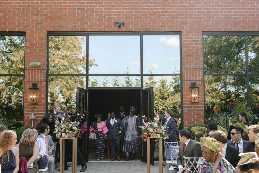 Guests gather outside a brick building with large windows for an outdoor event on a sunny autumn day.