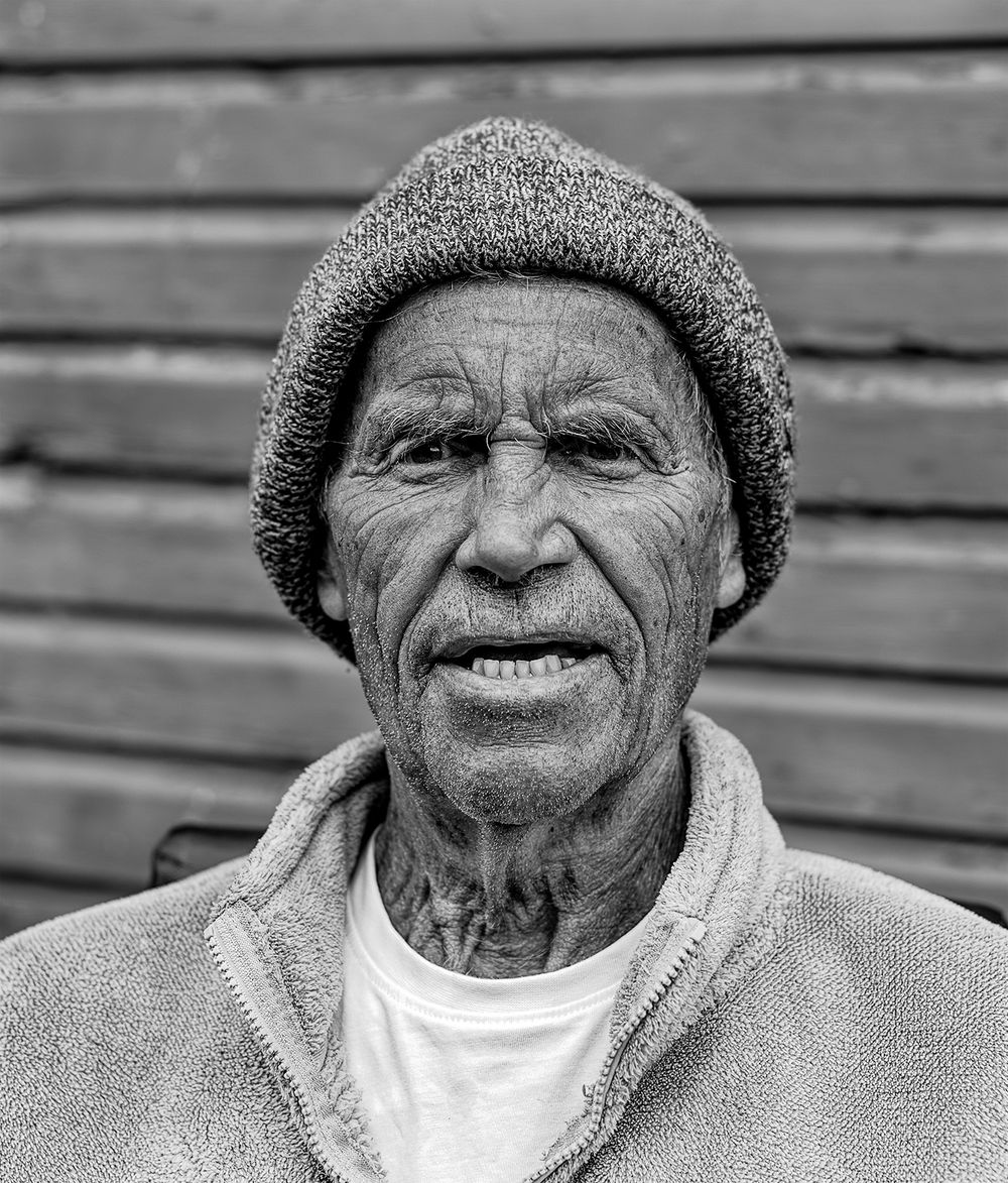 Black and white portrait photograph of an elderly person wearing a knit beanie and light colored sweater against wooden siding.