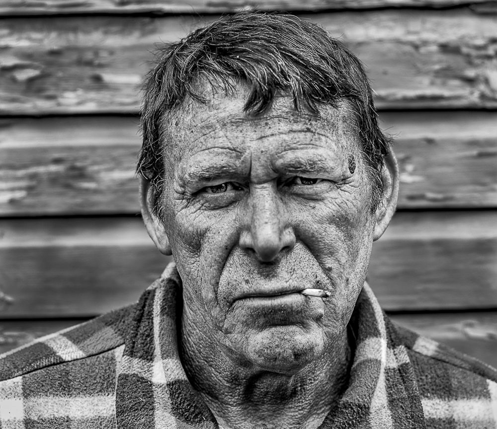Black and white portrait of a weathered person in a plaid shirt against rustic wooden siding.