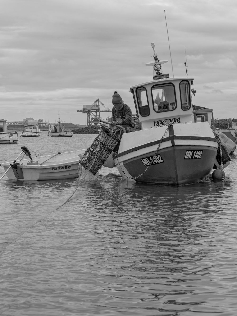 A small fishing boat tethered in harbor water with industrial cranes visible in the background under overcast skies.