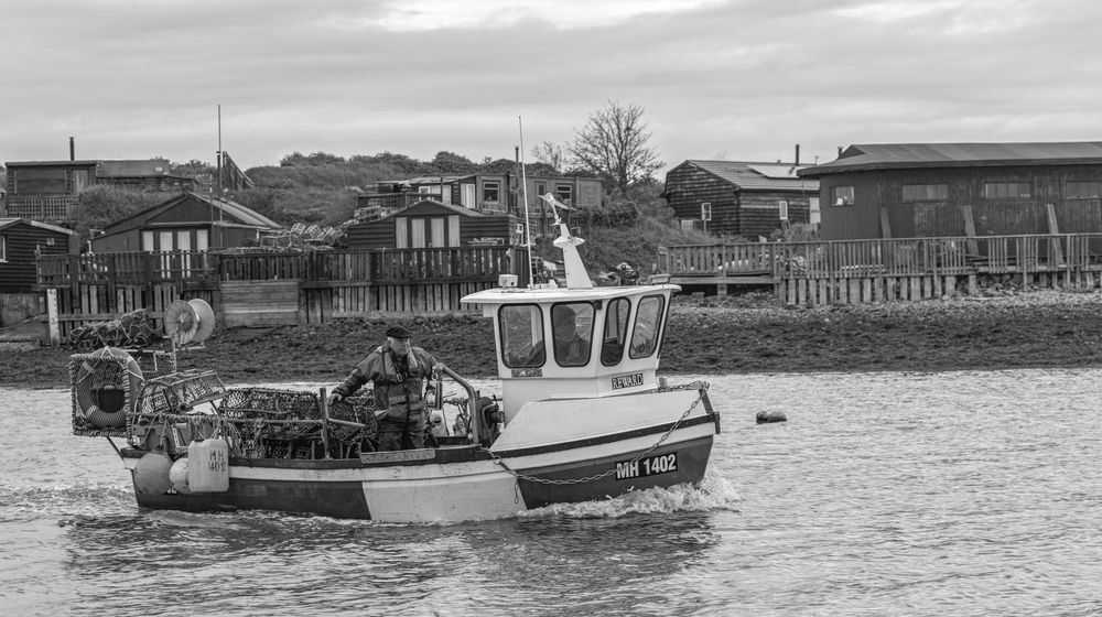 A small fishing boat navigates calm waters near a coastal town with houses and buildings visible on the shoreline.
